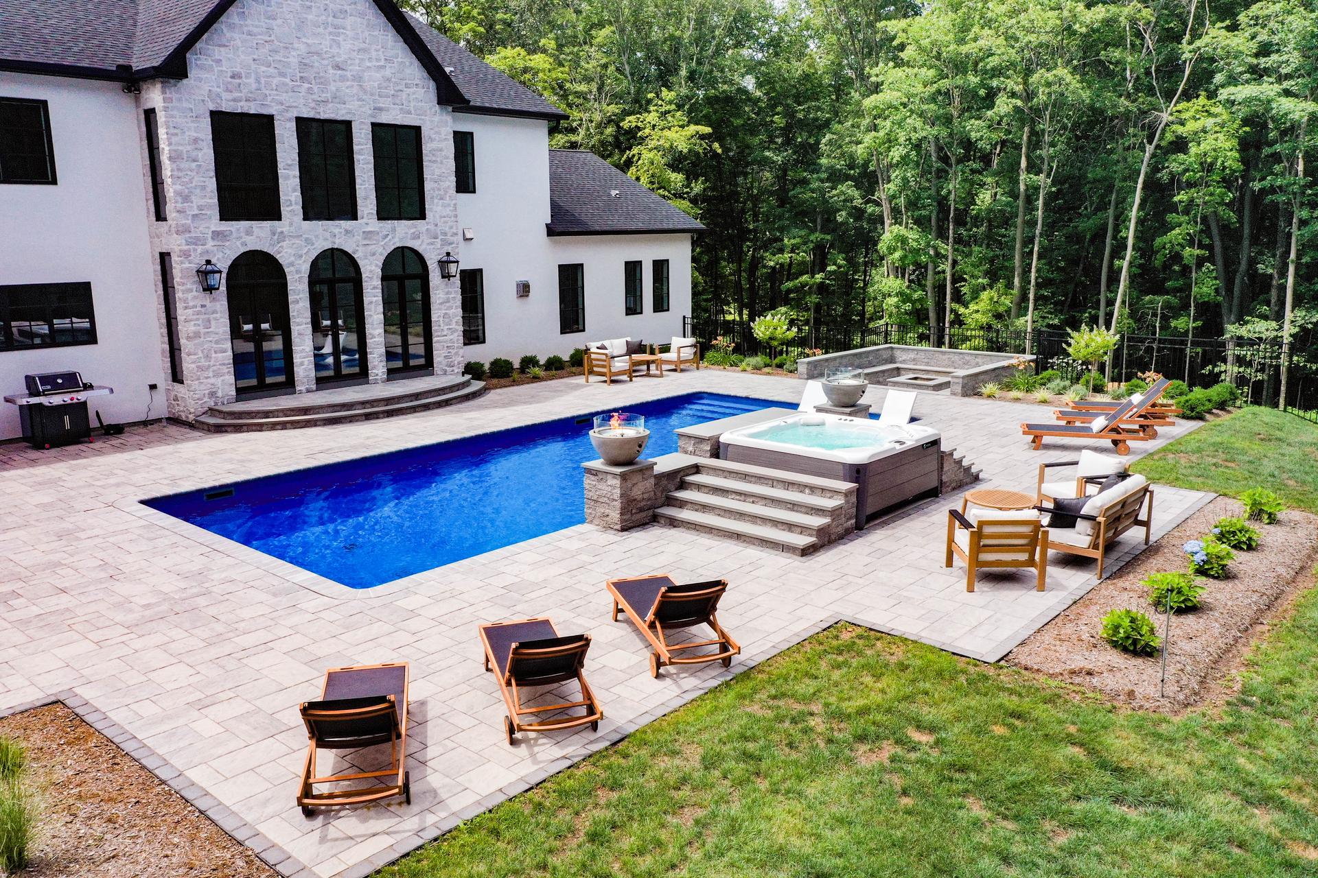 Custom pool patio layout in Connecticut featuring a large rectangular in-ground pool surrounded by light gray paver decking, with three wooden chaise lounges spaced along the pool edge, a raised spa with stone steps and fire bowls