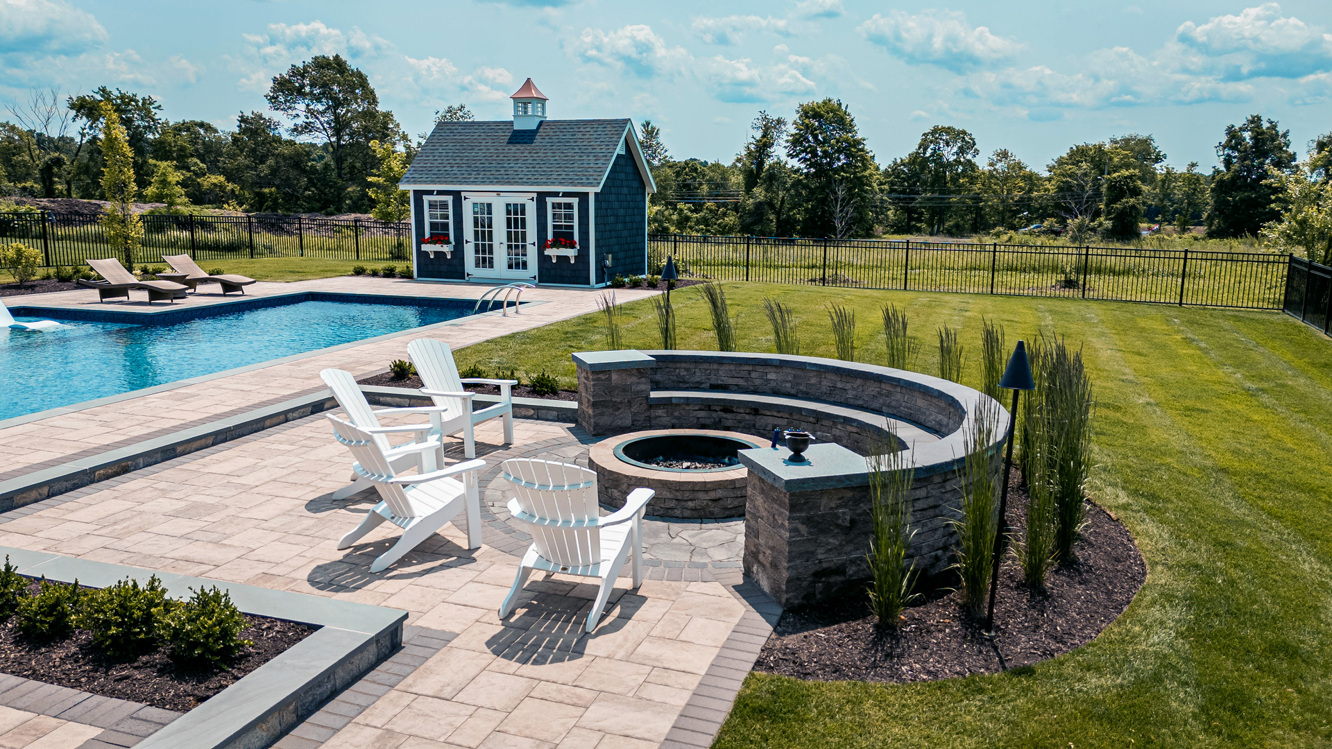 Custom Connecticut pool patio featuring a built-in stone fire pit with curved seating wall, paver patio, Adirondack chairs, swimming pool, and landscaped outdoor living space with ornamental grasses and a pool house in the background.
