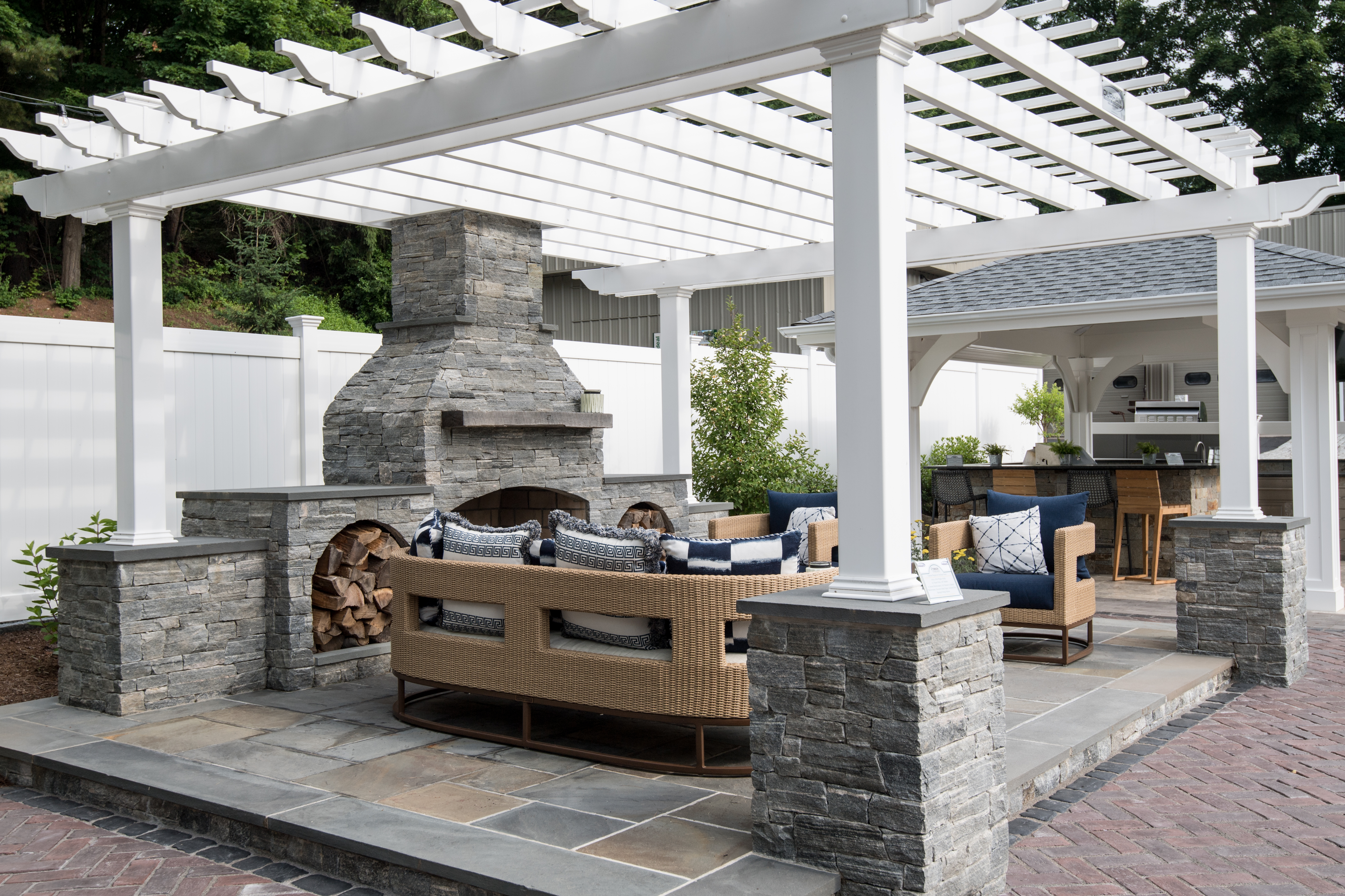 An image of a hardscape project completed by Torrison Stone and Garden.  A beautiful outdoor living space can be seen featuring a firepit, an l-shaped seating area with a table, a pillar-style heater for cooler months, a grilling area, a shade umbrella, a